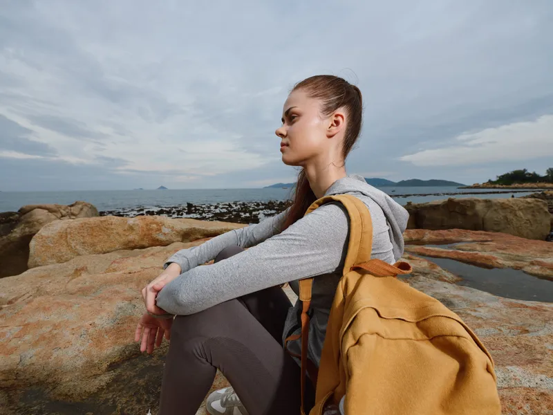 Young woman sitting on rocky beach, wearing casual gray hoodie and yellow backpack, contemplating the sea, showcasing emotions of peace and reflection in a natural setting
