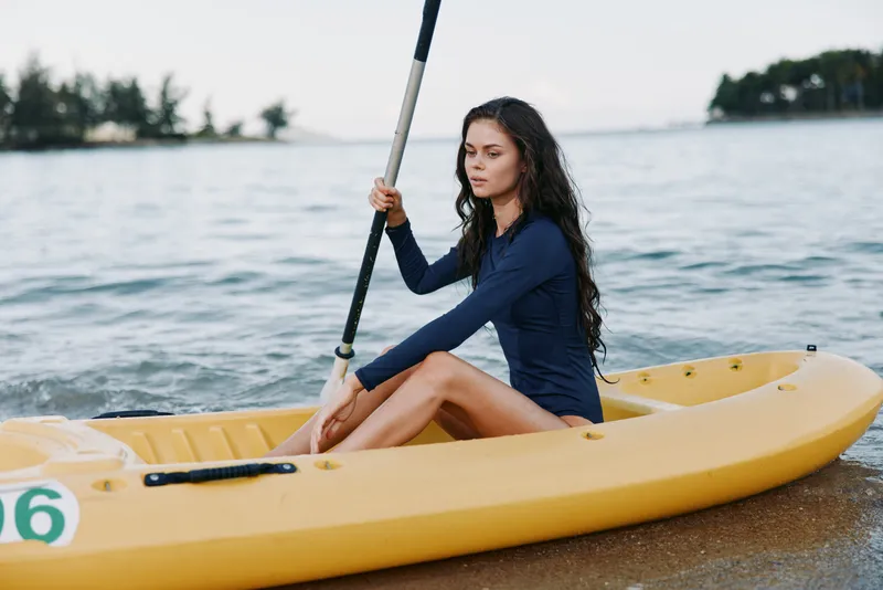 Young woman paddling yellow kayak on calm water during daytime, wearing navy blue outfit, concentrating and enjoying outdoor sport activity near shore.