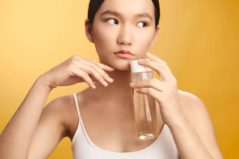 asian woman drinks water from a clear bottle, wearing a white tank top, as warm yellow background conveys a calm, healthy moment of hydration and focus.