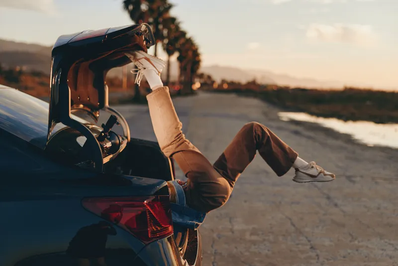 Woman outdoors by car trunk at sunset, travel lifestyle moment, relaxed casual pose, hands adjusting bag, warm golden light, road background, soft breeze, natural colors