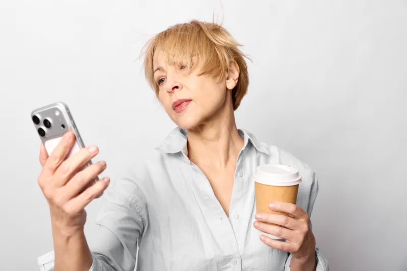 Woman holding smartphone and coffee cup in studio, casual white shirt, focused expression, morning routine, communication, copy space, soft light, isolated background