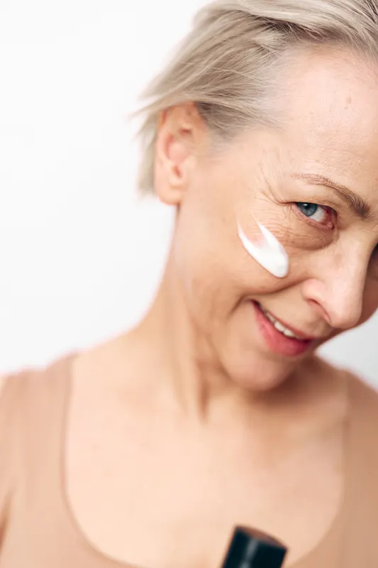 Mature woman with short hair applies white face cream on cheek, smiling gently. Concept of skin care, aging beauty, moisturizing, and natural wellness with soft natural lighting.