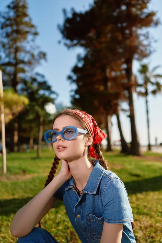 Lifestyle woman with modern boho-western denim street style wearing blue sunglasses and red bandana, sitting outdoors in warm film color lighting.