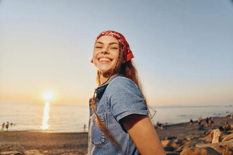 lifestyle woman smiling in modern boho-western denim street style with a red bandana, enjoying golden hour sunset by the beach in warm film color tones