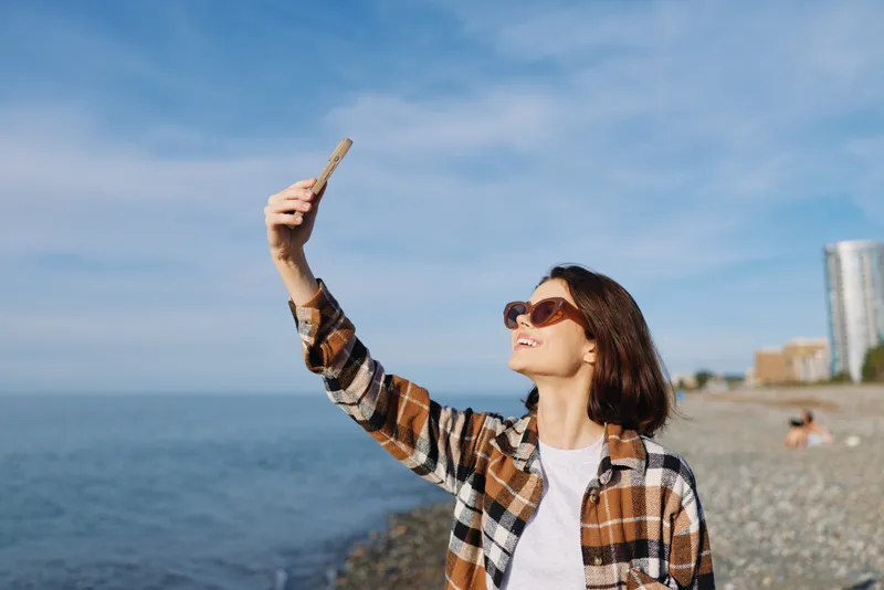 Selfie of a woman on a sunny beach wearing a plaid shirt and sunglasses, posing with a smartphone against a bright sky and calm blue sea.