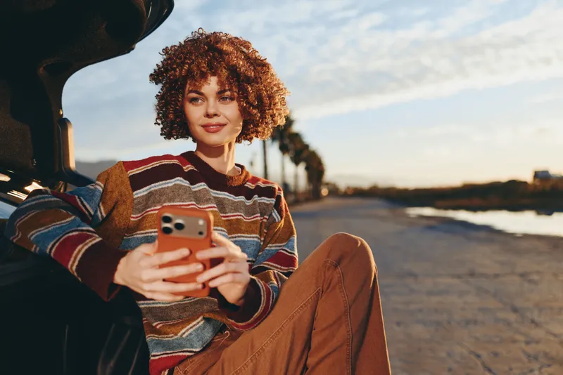 Woman lifestyle, smartphone in hand, curly hair, outdoor road, sitting by car at sunset, relaxed smile, casual sweater, warm evening light, natural colors, golden glow, distant landscape