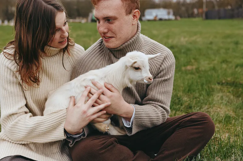 Young couple sitting on grass holds a small white baby goat together enjoying a peaceful moment in casual sweaters during an outdoor lifestyle scene.