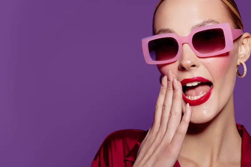 Stylish woman in pink sunglasses and red lipstick poses with hand near mouth on purple background. Concept of fashion, beauty, glamour, and confident expression.