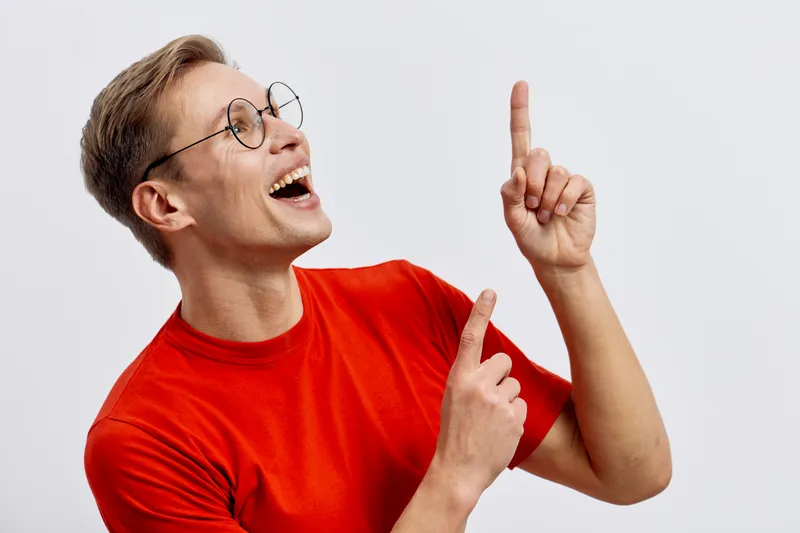 Cheerful young caucasian man wear red tshirt glasses pointing up smiling look side isolated on light grey background casual digital detox concept