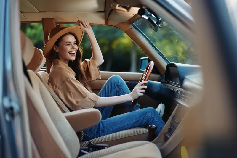 Happy young woman enjoying a road trip in a vintage car, dressed in a casual outfit with a hat, surrounded by nature.