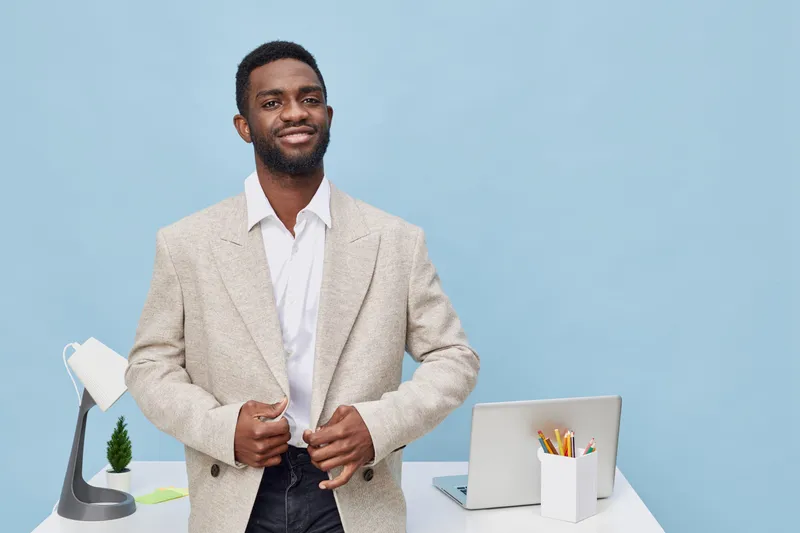 Confident smiling young African American man in beige blazer and white shirt standing at workspace isolated on light blue background. Modern professional business casual style for success and leadersh