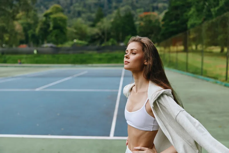 Confident young woman in sportswear enjoying fresh air on a tennis court, embodying fitness, wellness, and outdoor activity