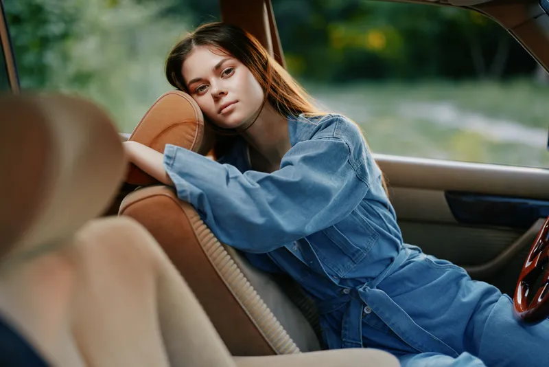 A young woman in denim jumpsuit relaxing in a car, showcasing a serene expression amidst a blurred outdoor backdrop, capturing a moment of tranquility.