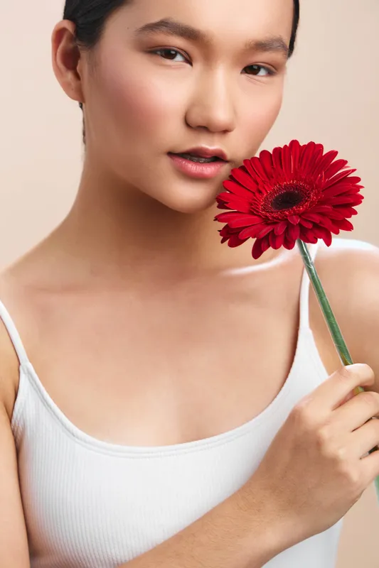 asian woman holds a vivid red gerbera flower close to her chest, eyes soft and expression calm, framed by bright studio light that highlights smooth skin and delicate features.