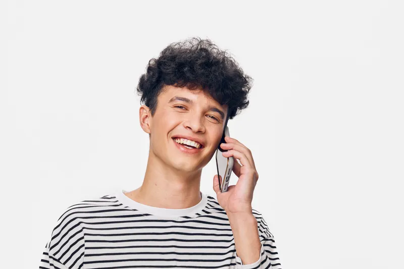 smiling man wearing striped shirt talking on mobile phone against white background, cheerful young person expressing happiness with curly hair