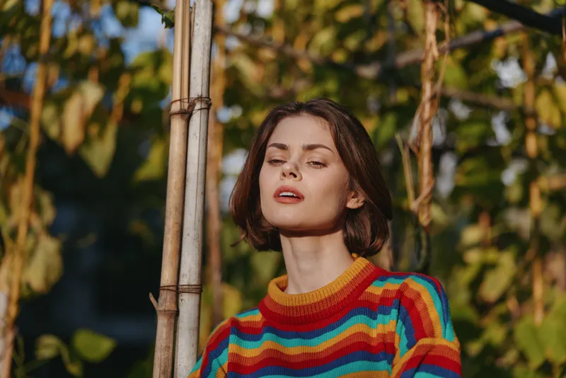 Woman in a colorful striped sweater with short brown hair poses in an outdoor portrait by a bamboo pole, warm sunlight on her face, confident expression, fashion lifestyle and autumn vibe