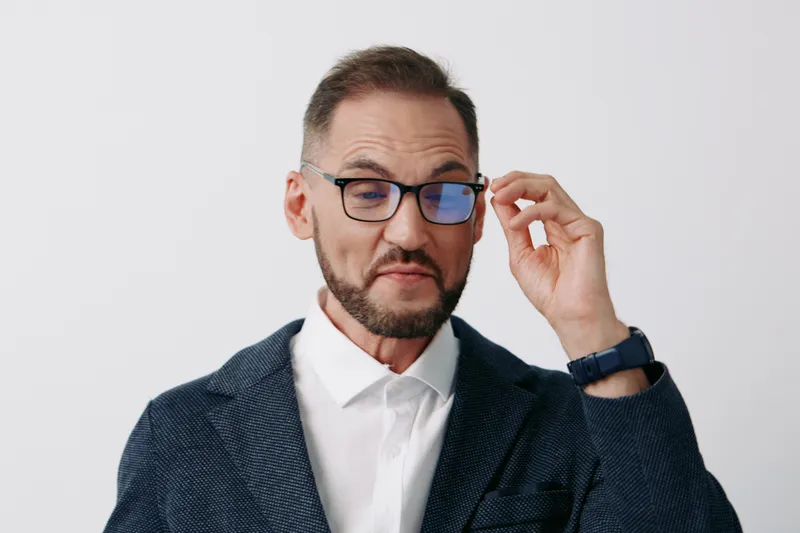 Businessman in a professional mood stands against a bright isolated colored background, adjusting glasses with a thoughtful gaze, conveying focus, control, and readiness.