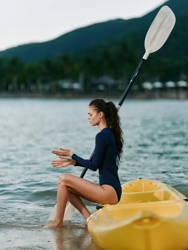 Young woman sits in shallow water beside a yellow kayak, adjusting a paddle. Sunlit beach, calm waves, and distant hills create a peaceful outdoor adventure scene.