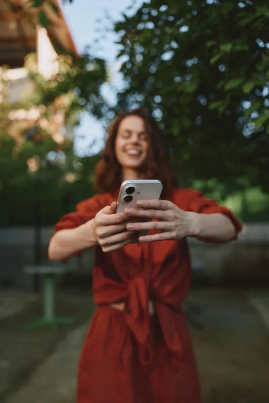 A smiling woman in a red robe enjoys a sunny outdoor moment, capturing a selfie with a smartphone. Bright greenery and a relaxed pose convey casual lifestyle and personal expression