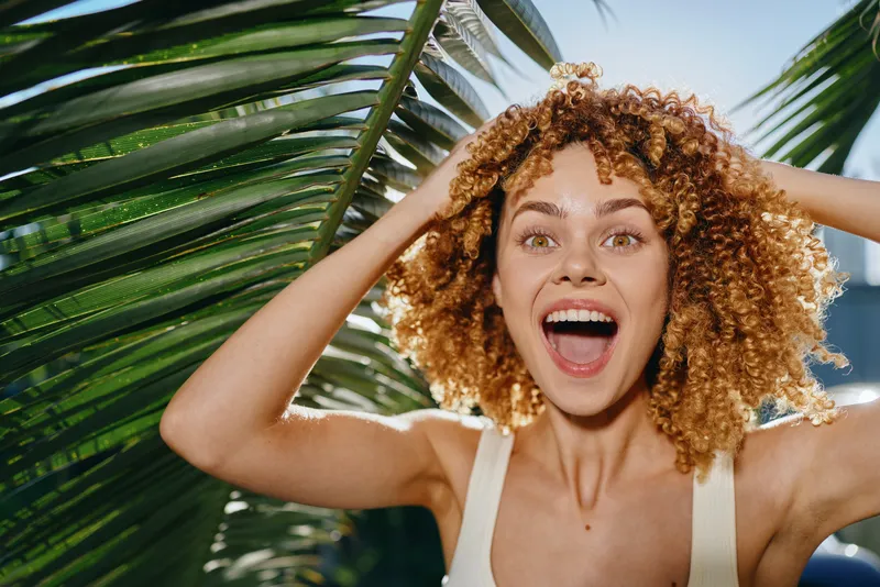 Excited woman with curly hair smiles brightly outdoors, surrounded by green tropical leaves, wearing a white tank top under natural sunlight and blue sky.