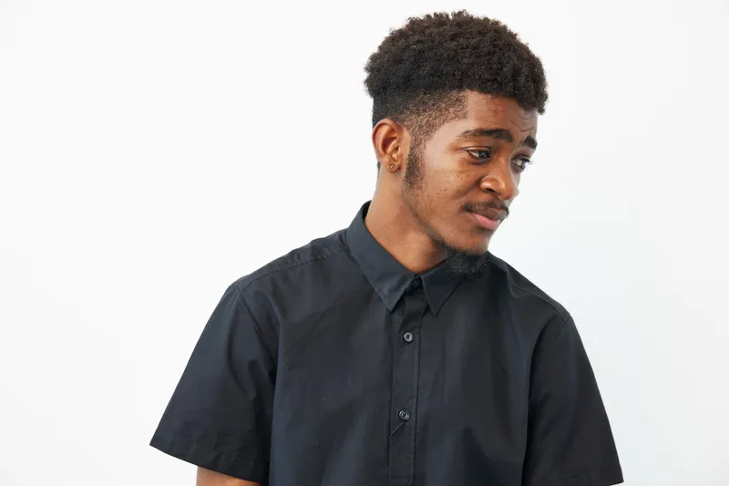 A thoughtful black man gazes to the side in a stylish black shirt, reflecting a moment of introspection and style, set against a minimalist background.