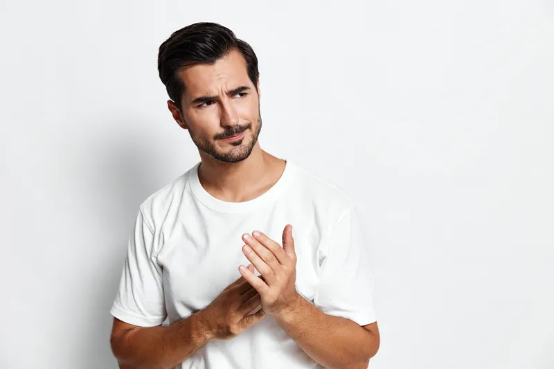 Thoughtful man with a questioning expression, wearing a plain white t-shirt, lightly touching his chest, isolated on a solid light background. Emotions and introspection theme.