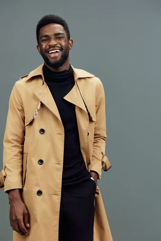Stylish young African man in a beige trench coat, smiling confidently against a muted gray background, perfect for fashion and lifestyle concepts