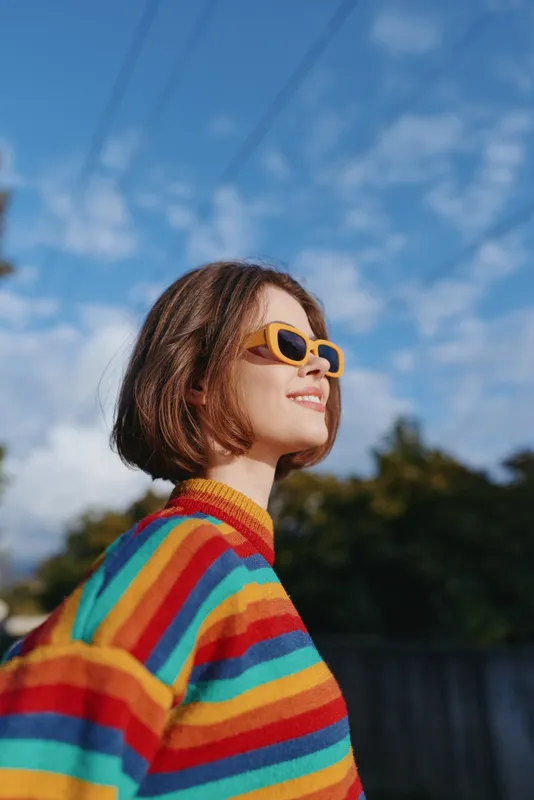 Woman smiling in a colorful striped sweater and yellow sunglasses, short hair bob, outdoors under blue sky. Casual lifestyle portrait, confident young adult enjoying a sunny day.
