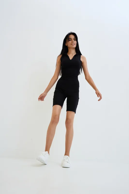 Slim woman wearing black sleeveless jumpsuit and white sneakers walking confidently against a light background in a minimalistic studio setting.
