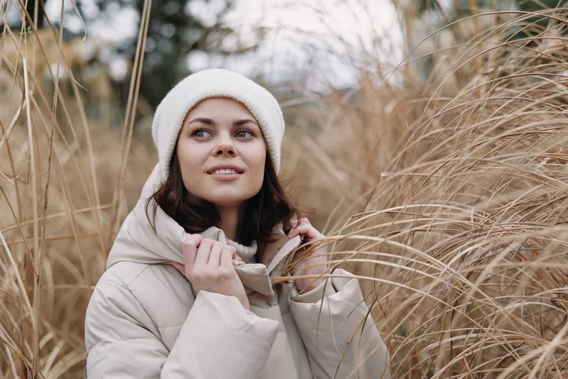 Woman outdoors field grasses wearing a beige winter coat and knit hat, a calm portrait in a natural setting with soft light and autumn mood in a open meadow