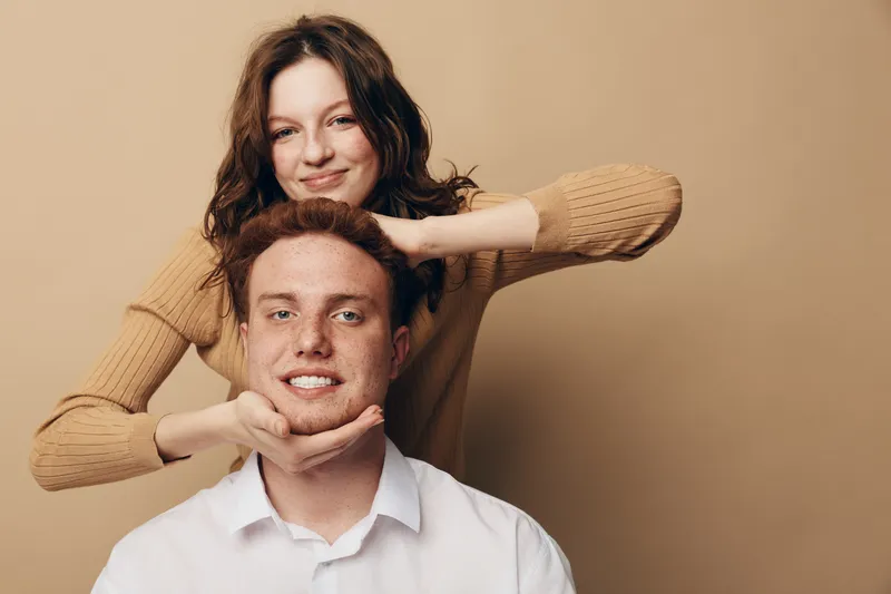 Smiling young woman in beige sweater gently holds head of happy redhead man in white shirt. Studio portrait on brown background. Concept of friendship, affection, and positive emotions.