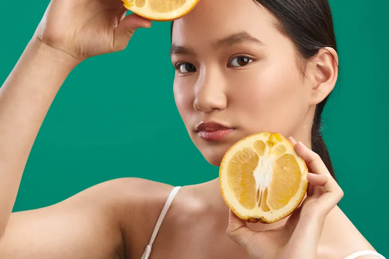 Asian woman holds citrus slices against a vivid studio backdrop, presenting a fresh healthy lifestyle moment. Her calm expression and soft lighting convey wellness and modern beauty in a bright