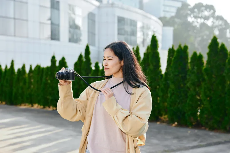 Camera in hand, woman photographs outdoors in an urban setting with a casual jacket, focused expression, modern building and greenery in the backdrop, a street style photo documenting daily