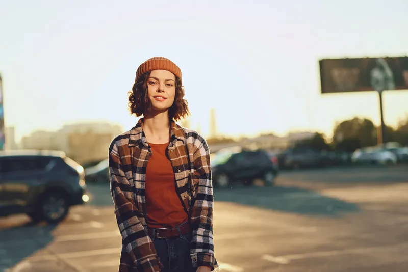 Woman portrait in golden hour urban parking lot, beanie and plaid jacket, casual smile and sunlight backlight. Authenticity, candid lifestyle and emotional storytelling vibe.