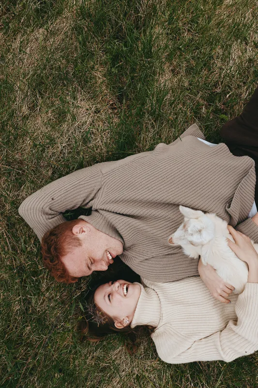 Couple lying on grass in cozy sweaters, smiling and enjoying a relaxed lifestyle, warm soft light, intimate connection, calm wholesome mood, outdoor park setting, natural tones, gentle breeze, pure