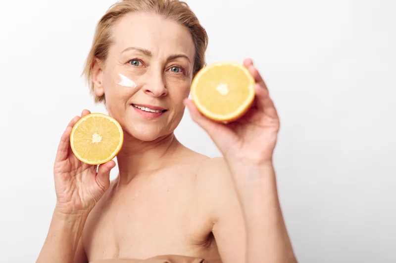 Smiling mature woman with short hair holding two halves of an orange near her face with cream on cheek. Healthy skin concept with natural skincare and vitamin C beauty treatment.
