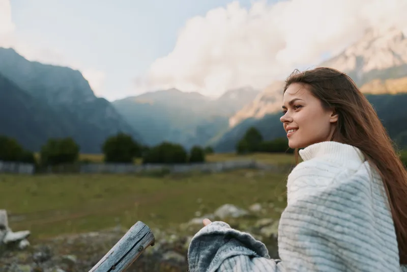 Woman sits outdoors in a cozy sweater overlooking mountain scenery, enjoying a peaceful lifestyle moment in nature, casual and serene in the countryside