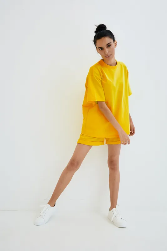 Young woman wearing bright yellow casual outfit poses confidently against white background, showcasing modern minimalistic style and sporty footwear in natural light.
