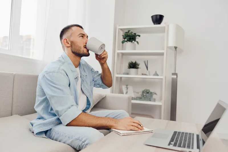 Young man enjoying coffee while working remotely in a modern living room, focusing on a laptop and taking notes, creating a productive atmosphere.
