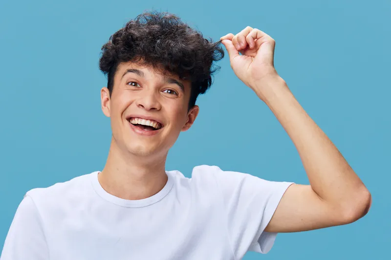 Happy young man with curly hair wearing white t-shirt smiling brightly against a blue background, expressing joy and positive energy in casual style.