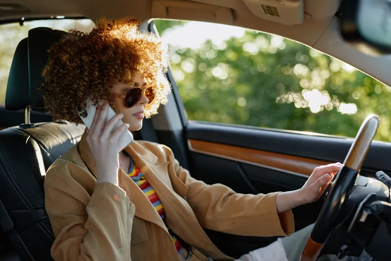 Young woman with curly hair drives a car, talking on a smartphone, wearing sunglasses and a tan blazer, capturing a retro film vibe with warm light and fashionable confidence