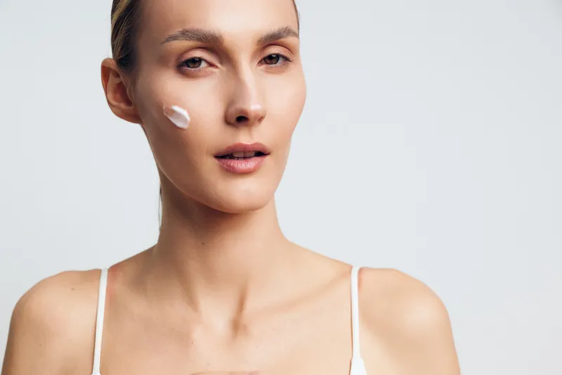 Close up of woman with natural skin applying face cream on cheek, soft lighting, minimal makeup, clean background, skincare routine concept, calm expression
