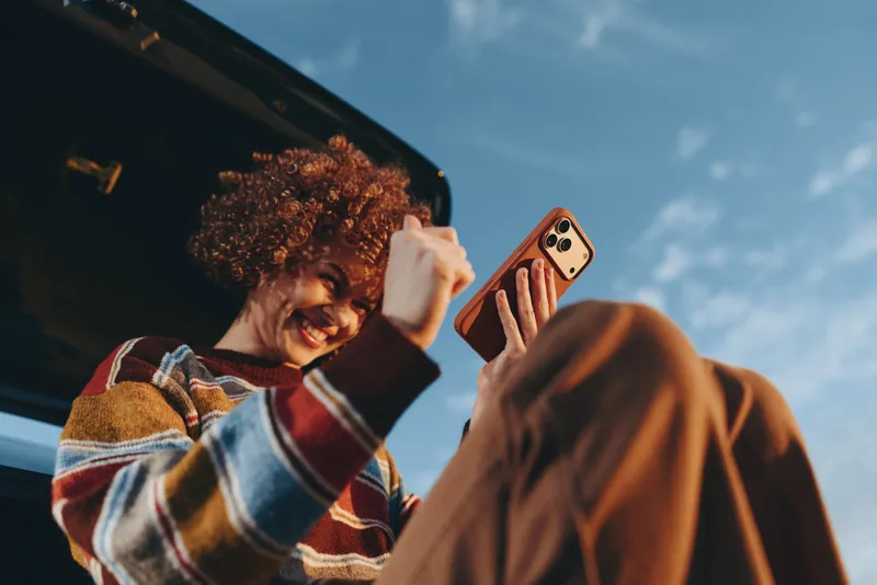 Woman lifestyle selfie outdoors near a car, curly haired woman holds smartphone and smiles toward the camera, sunlight and blue sky create a relaxed casual travel mood.