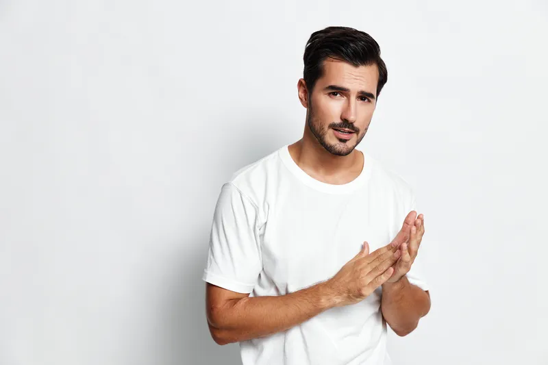 Handsome young man in a casual white t-shirt looking thoughtfully at the camera with a slight smile, standing against a plain light background. People lifestyle concept.