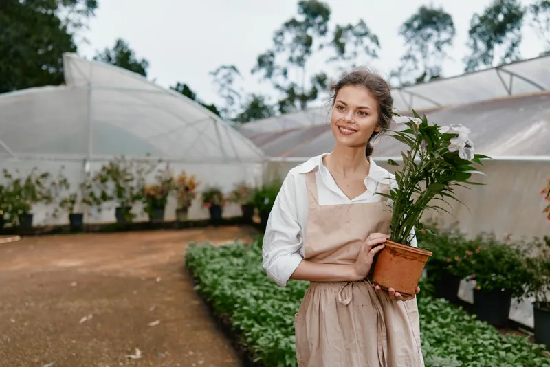 Young woman in a greenhouse holding a potted plant, smiling and dressed in a casual outfit, embodies joy, gardening, and greenery in a serene setting