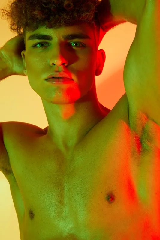 Young man with curly hair posing shirtless with hands behind head under colorful red and green lighting. Closeup portrait with intense gaze and smooth skin texture in studio.