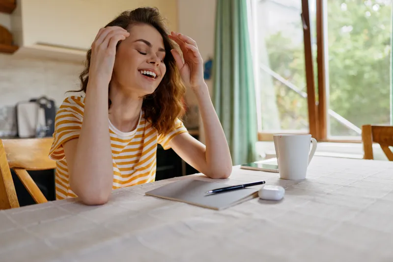 Smiling woman with striped shirt sits at a home table, writing in a notebook. Natural daylight from a window highlights a cozy, casual lifestyle moment.