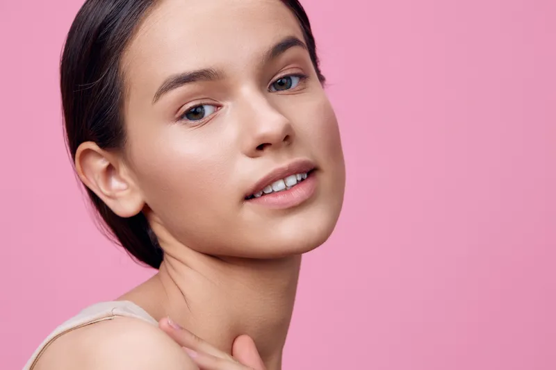 Young woman with natural beauty and soft smile against a pastel pink background, showcasing healthy skin and a minimalistic style