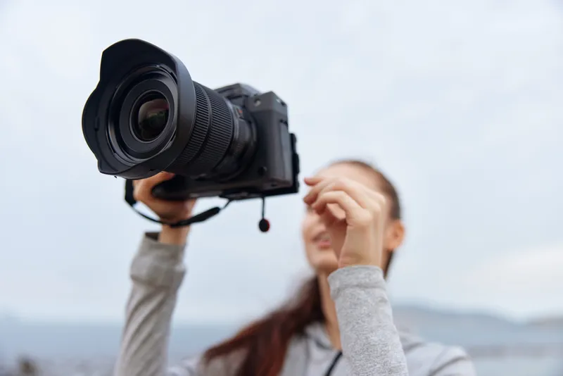 Young woman with a camera smiles outdoors, capturing moments, showcasing enthusiasm for photography, with a cloudy background emphasizing creativity and passion