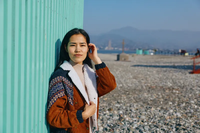 Woman leaning against a turquoise wall on a pebble beach, wearing a knit coat and scarf, a calm coastal scene with natural light and outdoor charm, candid mood